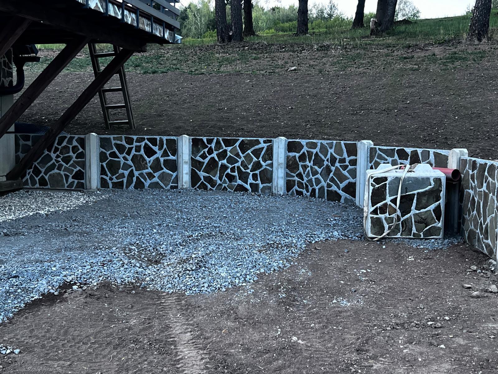 Betónový oporný múr s kamennou úpravou počas dokončovania, pred ním štrková plocha